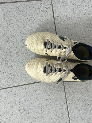 Vita Nike fotbollsskor med svarta detaljer tiempos - Säljer ett par vita fotbollsskor från Nike med svarta detaljer och snörning. Skorna har en klassisk låg profil och är tillverkade i syntetmaterial med vadderad insida för extra komfort. Perfekta för dig som vill ha stilrena och funktionella skor på planen.