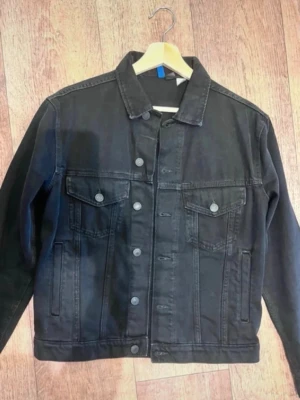 Svart jeansjacka med tryck bak - Snygg svart jeansjacka med klassisk passform och två bröstfickor med knappar. Baksidan har ett stort tryck med motiv i vitt. Perfekt för dig som vill ha en unik och trendig look. Knappstängning framtill och raka ärmar.