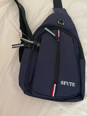 Mörkblå axelväska från SFUTE - Snygg mörkblå axelväska från SFUTE med justerbar svart rem och flera dragkedjefack. Väskan har en vit logga, röd-vit-blå randig detalj framtill och är tillverkad i slitstarkt syntetmaterial. Perfekt för dig som vill ha en sportig och praktisk look.