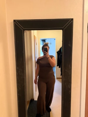 Brun off shoulder jumpsuit - Säljer en brun jumpsuit med off shoulder-design och vida ben. Jumpsuiten har kort ärm och sitter figurnära upptill, vilket ger en snygg siluett. Perfekt för dig som gillar en enkel men trendig look. Materialet ser ut att vara mjukt och stretchigt för extra komfort.