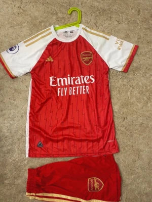 Arsenal matchställ Adidas röd/vit - Säljer ett Arsenal matchställ från Adidas med röd tröja och vita ärmar, guldiga detaljer och klubbmärke på bröstet. Tröjan har trycket 'Emirates Fly Better' och spelarnamn G.Jesus med nummer 9 på ryggen. Tillhörande röda shorts med klubbmärke och gulddetaljer.