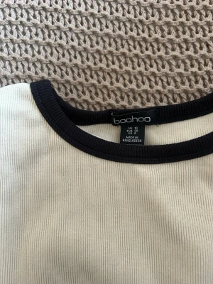 Ribbad vit t-shirt från Boohoo - Snygg ribbad vit t-shirt från Boohoo med svarta kontrastkanter vid halsringning och ärmslut. T-shirten har en klassisk passform och är kortärmad, perfekt till jeans eller kjol.. Aldrig använd.