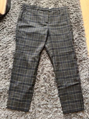 Rutiga kostymbyxor i brunt - Snygga kostymbyxor med rutigt mönster i brunt och mörkblått. Byxorna har raka ben, bälteshällor och sidofickor. Perfekta för en stilren och trendig look.