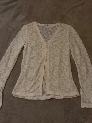 Vit spetskofta med pärlknappar - Säljer en tunn, vit spetskofta från NA-KD med v-ringning och långa ärmar. Koftan har dekorativa pärlknappar framtill och är gjord i ett genomskinligt spetsmaterial som ger en romantisk vibe. Perfekt att styla över ett linne eller klänning.