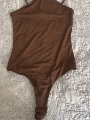 Brun body med smala axelband - Säljer en stilren brun body från H&M med smala axelband och åtsittande passform. Tillverkad i ett mjukt och stretchigt material som ger en snygg siluett. Perfekt att matcha med jeans eller kjol för en trendig look.