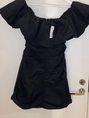 Svart offshoulder klänning Lindex - Trendig svart kortklänning från Lindex i återvunnen bomullsblandning. Klänningen har offshoulder-modell med puffiga ärmar och en smickrande passform. Perfekt för dig som vill ha en stilren och modern look.