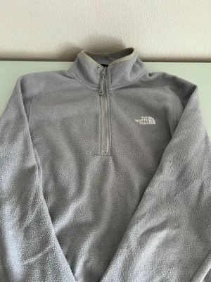 Grå fleece från The North Face - Mysig grå fleece från The North Face med halv dragkedja och hög krage. Tröjan har broderad logga på bröstet och är tillverkad i mjukt fleecematerial. Perfekt för lager-på-lager och håller dig varm under kyliga dagar.