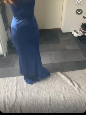 Marinblå långklänning med snörning - Säljer en marinblå långklänning i glansigt material med smala axelband och snygg snörning i ryggen. Klänningen har en figurnära passform och faller elegant längs kroppen. Perfekt för dig som vill sticka ut på nästa event! Skriv för mer bilder