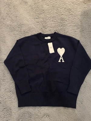 Mörkblå stickad tröja från Ami - Snygg mörkblå stickad tröja från Ami Alexandre Mattiussi med ikonisk vit hjärta-A logga på bröstet. Tröjan har lång ärm, ribbade muddar och rund hals. Perfekt för dig som gillar stilrena detaljer och vill ha något som sticker ut.