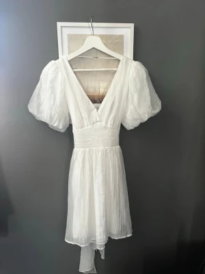 Vit klänning - Superfin vit kortklänning med omlott-design och stora puffärmar. Klänningen har en v-ringad fram och bak samt en stor rosett baktill. Materialet är lätt och plisserat, vilket ger en luftig känsla. Perfekt för sommarens alla tillfällen. Stl 34, passar mig som är xs. Ny pris 699kr säljer för 525
