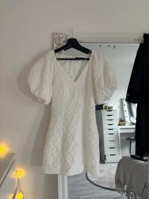 Vit kort klänning med puffärmar - Trendig vit kortklänning med v-ringning och puffiga kortärmar. Klänningen har ett subtilt strukturmönster i tyget och en feminin siluett. Perfekt för sommarens alla tillfällen och lätt att styla med accessoarer.