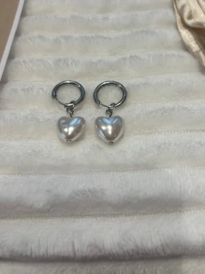 Silverörhängen med pärlhjärtan - Säljer ett par stilrena örhängen i silverfärg med hjärtformade pärlor som hänger i små ringar. Perfekta för dig som gillar en söt och trendig look. Hjärtana har en skimrande pärlfinish och örhängena är lätta att bära.
