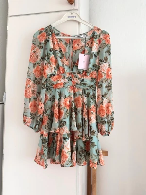 Blommig kortklänning från Hello Molly - Supersöt kortklänning från Hello Molly med blommigt mönster i orange, grön och vit på ljus botten. Klänningen har v-ringning, långa ärmar med resår och volangdetaljer i kjolen. Perfekt för sommarens alla tillfällen!