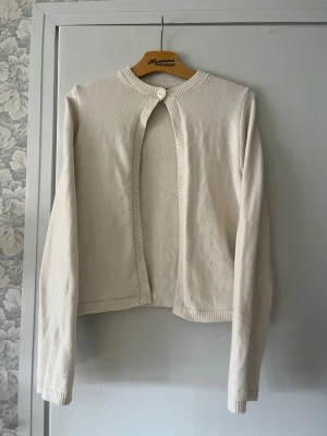 Beige kofta från Gina Tricot - Stilren beige kofta från Gina Tricot med rund halsringning och en knapp framtill. Koftan har långa ärmar och ribbade muddar vid ärmslut och nederkant. Perfekt att slänga över en topp för en enkel och clean look.