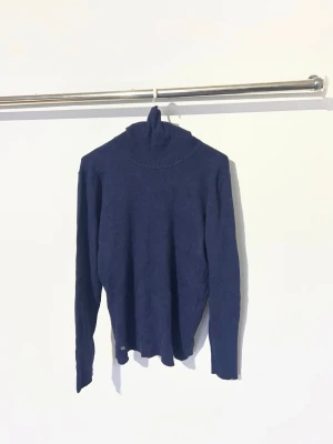 Mörkblå polotröja från Ralph Lauren - Snygg mörkblå stickad polotröja från Ralph Lauren med lång ärm och ribbade muddar. Tröjan har en klassisk polokrage och en liten metallbricka med RL-logga vid nederkanten. Perfekt för lager på lager och stilrena outfits.