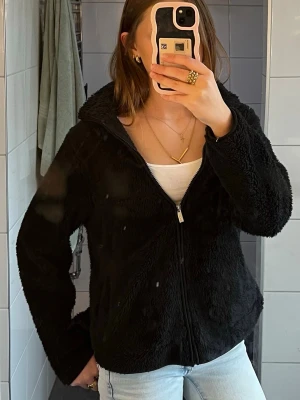 Svart teddyjacka med dragkedja - Mysig svart teddyjacka med dragkedja framtill och hög krage. Jackan har en avslappnad passform och är gjord i fluffigt, mjukt material som ger en cool och trendig look. Perfekt att slänga på sig över en hoodie eller t-shirt för extra värme.