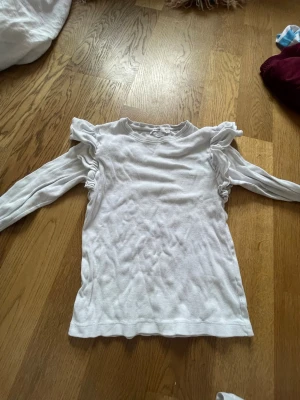 Vit långärmad topp med volang - Snygg vit långärmad topp med volangdetaljer vid axlarna som ger en trendig look. Tillverkad i mjukt bomullsmaterial och har en normal passform. Perfekt att styla med jeans eller kjol för en clean och fräsch outfit.