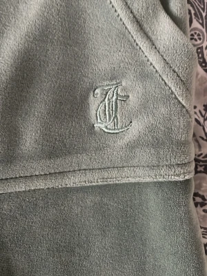 Gröna mjukisbyxor från Juicy Couture - Snygga gröna mjukisbyxor från Juicy Couture i mjuk velour. Byxorna har raka ben, två bakfickor med knappar och broderad JC-logga framtill. Perfekta för en chill och trendig look. Byxorna har en knappt synlig brun färg längst ner vid fållen längst ner. Byxornas material är i nyskick och är väldigt bekvämt. Byxorna är även upplagda och passar någon som är runt 160 cm lång