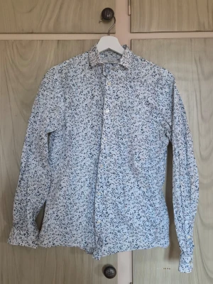 Vit bladig skjorta med blå och röda detaljer - Snygg vit skjorta med småblqdigt mönster i blått och rött. Klassisk krage, knäppning framtill och långa ärmar. Perfekt för dig som vill ha en fräsch och stilren look med lite extra detaljer. Tillverkad i mjuk bomull för skön känsla. Alla kläder rollas innan dom skickas hem då det finns katt i hemmet🐈 Jag stryker och tvättar INTE innan jag skickar det får du stå för själv så det blir som du vill ha det🤗