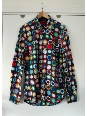 Desigual - färgglad mönstrad skjorta - storlek S - Unik skjorta från Desigual i storlek S med ett färgglatt mönster av cirklar i olika storlekar och färger. Skjortan har långa ärmar med manschetter och en klassisk krage. Knapparna är mörkbruna med Desigual-logotypen ingraverad. En röd Desigual-logotyp finns broderad på baksidan av kragen. 100% bomull, så bra kvalitativt material som andas. Nypris 1359:-. Mycket gott skick, som ny!