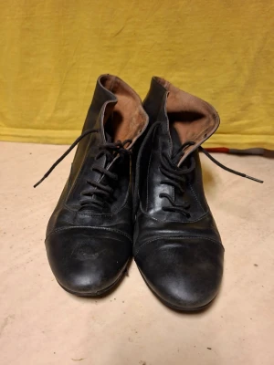 Svarta snörkängor i läder - Klassiska svarta snörkängor i äkta läder med snörning framtill och låg klack. Skorna har en elegant, smal siluett och rundad tå. Insidan är fodrad i brunt tyg för extra komfort. Perfekta för dig som gillar tidlös och stilren design.