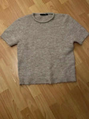 Beige stickad topp kort ärm - Mysig beige stickad topp med korta ärmar och rund hals. Perfekt för lager på lager och enkel att matcha med olika stilar. Ribbad kant nedtill och vid ärmslut för en snygg finish.