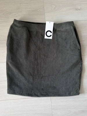 Mörkgrön manchesterkjol från C - Snygg mörkgrön kortkjol i manchester från C. Kjolen har raka linjer, två sidofickor och resår i midjan för en bekväm passform. Perfekt att matcha med en stickad tröja eller t-shirt för en cool look.