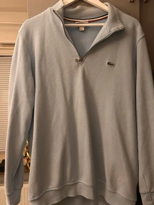 Ljusblå half zip tröja från Lacoste - Säljer en ljusblå långärmad tröja från Lacoste med half zip och klassisk krokodillogga på bröstet. Tröjan har en stilren design och är tillverkad i bomull, perfekt för lager-på-lager under kyliga dagar. Finns en jätte liten defekt på den men syns knappt. Passar M också!