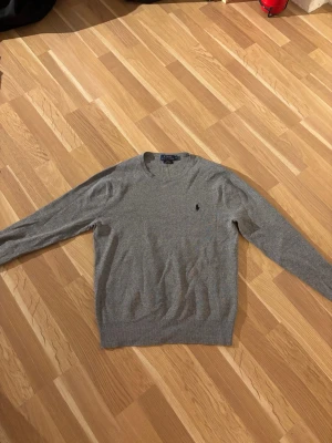 Grå stickad tröja Polo Ralph Lauren - Snygg grå stickad tröja från Polo Ralph Lauren i slim fit-modell. Tillverkad av mjuk pima cotton och har rund halsringning samt den klassiska lilla loggan broderad på bröstet. Perfekt till jeans eller chinos för en clean look.