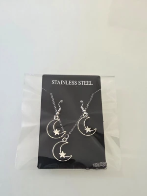 Smyckeset i rostfritt stål med måne - Snyggt smyckeset i rostfritt stål med halsband och örhängen. Både halsbandet och örhängena har hängsmycken i form av en måne med en liten stjärna. Kedjan är tunn och silvrig, och smyckena har en modern och stilren look. Tål vatten och missfärgas inte eftersom det är i rostfritt stål! Perfekt som present. 