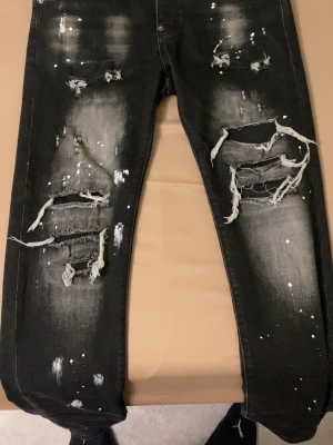 Svarta slitna jeans med färgstänk - Säljer ett par svarta jeans med kraftiga slitningar och trasiga detaljer framtill. Jeansen har vita färgstänk och en tvättad look som ger en riktigt edgy vibe. Perfekta för dig som gillar streetwear och vill sticka ut med din stil.