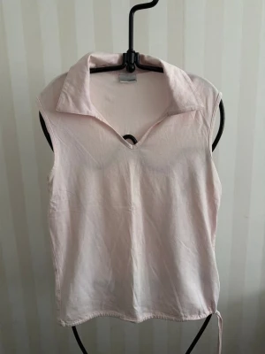 Casall Ljusrosa ärmlös topp med krage storlek M ny - Snygg ljusrosa ärmlös topp med krage och v-ringning. Toppen har en enkel och clean design med knyt i sidan för extra detalj. Perfekt till jeans eller kjol för en chill look.