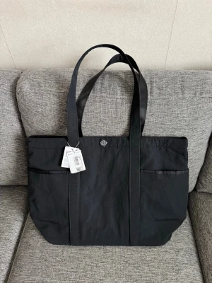 Svart totebag  från Lululemon - Stilren svart shoppingväska från Lululemon i slitstarkt nylonmaterial. Väskan har dubbla axelband, flera ytterfickor och en diskret logga framtill. Perfekt för dig som vill ha en rymlig och praktisk tote med enkel design.