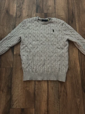Ljusgrå kabelstickad tröja Polo Ralph Lauren - Snygg ljusgrå kabelstickad tröja från Polo Ralph Lauren med rund halsringning och svart broderad logga på bröstet. Tröjan har ribbade muddar vid ärmslut och nederkant och är perfekt för dig som gillar klassisk stil.