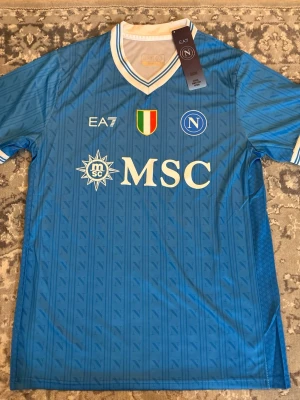 Napoli matchtröja blå EA7 - Snygg blå Napoli matchtröja från EA7 med vita detaljer och klubbmärke på bröstet. Tröjan har V-ringning, tryckt sponsorlogga MSC och italienska mästarmärket. Perfekt för fotbollsfans som vill visa sitt stöd för laget.