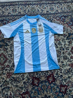 Argentina matchtröja Adidas blåvit - Säljer en officiell Argentina matchtröja från Adidas i klassiska blå och vita ränder. Tröjan har guldiga detaljer, FIFA-märke och AFA-logga på bröstet samt tre blå ränder på axlarna. Tillverkad i lätt och ventilerande material, perfekt för fotboll.