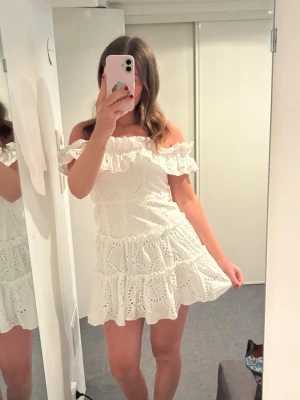 Vit offshoulder klänning med volang - Söt vit klänning med offshoulder och volangdetaljer. Klänningen har broderade hålmönster och en luftig, kort passform med flera lager. Perfekt för sommarens alla tillfällen och ger en romantisk vibe.