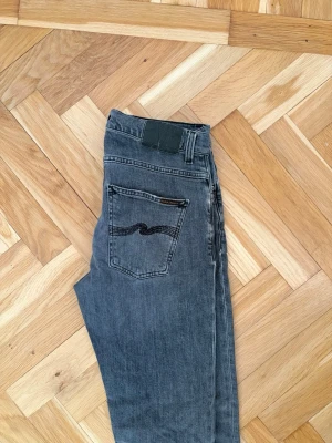 Grå jeans från Nudie Jeans - Säljer ett par grå jeans från Nudie Jeans med klassisk femficksdesign och svarta sömmar på bakfickan. Jeansen har rak passform och är tillverkade i denim med en lätt tvättad look. Perfekta för en avslappnad stil.