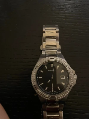Silverfärgad klocka från GANT - Stilren klocka från GANT i silverfärgat metallarmband och svart urtavla. Urtavlan har datumvisning och är omgiven av en glittrig kant med stenar. Perfekt accessoar för att lyfta din outfit.