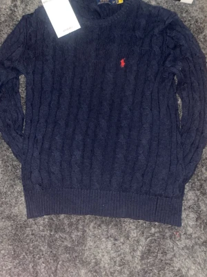 Mörkblå kabelstickad tröja Polo Ralph Lauren - Mörkblå kabelstickad tröja från Polo Ralph Lauren med klassisk röd broderad logga på bröstet. Tröjan har rund halsringning, långa ärmar och ribbstickade muddar. Perfekt för en stilren och tidlös look.