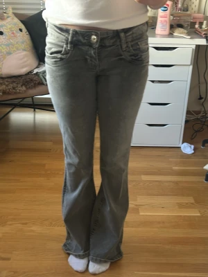 Grå bootcut jeans med låg midja - Säljer ett par grå bootcut jeans med låg midja och klassiska femficksdetaljer. Jeansen har en snygg tvättad look och vida ben nertill. Perfekta för dig som gillar retrostil och vill ha ett par bekväma jeans med cool attityd.