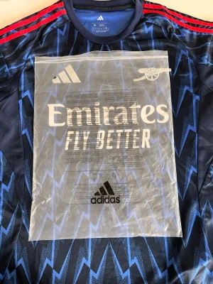 Arsenal bortatröja blå Adidas - Snygg Arsenal bortatröja från Adidas i mörkblått med blått blixtmönster och röda ränder på axlarna. Tröjan har korta ärmar, rund hals och tryck med Emirates Fly Better på bröstet. Perfekt för fotbollsträning eller att bära som supporter.