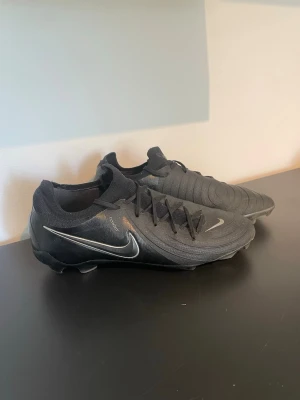 Svarta Nike fotbollsskor Phantom GX - Snygga svarta Nike Phantom GX fotbollsskor med låg profil och strumpliknande skaft. Skorna har klassisk snörning, diskret silverfärgad Nike-swoosh och är tillverkade i syntetmaterial för bra grepp och kontroll på planen.