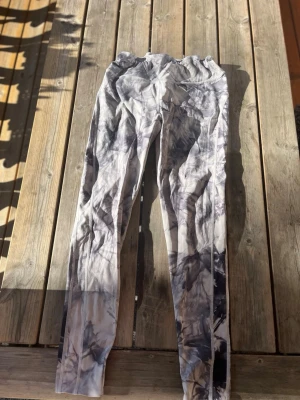 Grå-svarta tie dye träningstights - Snygga träningstights med grå och svarta tie dye-mönster. De är långa och har en tight passform, perfekta för gymmet eller löpning. Materialet känns stretchigt och följsamt, vilket ger bra rörelsefrihet under träning. Har två knutar vid midjan, en liten brun fläck som man kan se på fjärde bilden. Men annars superfina men kommer inte till användning. Det står inte vilken storlek men skulle gissa på S/M eftersom jag själv har det och dem passar.