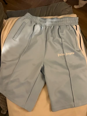 Palm Angels ljusblå shorts - Säljer ett par ljusblå shorts från Palm Angels med vita och svarta ränder längs sidorna. De har elastisk midja, raka ben och Palm Angels-loggan broderad i vitt framtill. Dom har en liten dock ganska diskret fläck (se bilden)