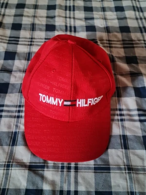 Röd keps från Tommy Hilfiger - Snygg röd keps från Tommy Hilfiger med broderad logga framtill och diskret mönster av märkesnamnet över hela kepsen. Klassisk böjd skärm och justerbar baksida. Perfekt för en sportig och trendig look.
