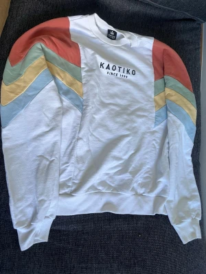 Vit sweatshirt från Kaotiko - Vit sweatshirt från Kaotiko med trycket 'KAOTIKO SINCE 1999' på bröstet. Ärmarna har färgglada ränder i blått, gult, grönt och rött som ger en retro känsla. Rund halsringning och ribbade muddar. Perfekt för en avslappnad stil.
