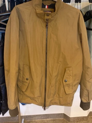 Beige vindjacka från Tommy Hilfiger - Stilren beige vindjacka från Tommy Hilfiger med hög krage, dragkedja framtill och två fickor med lock och tryckknapp. Jackan har ribbade muddar vid ärmslut och nederkant samt diskret logodetalj i nacken. Perfekt för blåsiga dagar.