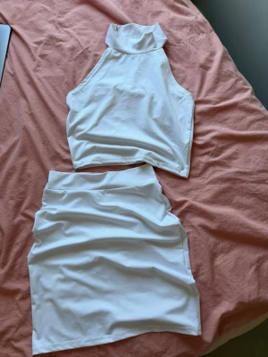Vitt tvådelat set med hög krage - Säljer ett vitt tvådelat set med crop top och hög krage samt matchande kjol. Setet har en stilren och enkel design i mjukt stretchigt tyg, perfekt för en trendig look.