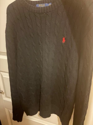 Svart kabelstickad tröja Polo Ralph Lauren - Klassisk svart kabelstickad tröja från Polo Ralph Lauren med rund halsringning och röd broderad logga på bröstet. Tröjan har långa ärmar och ribbade muddar. Perfekt för dig som gillar tidlös och stilren design.